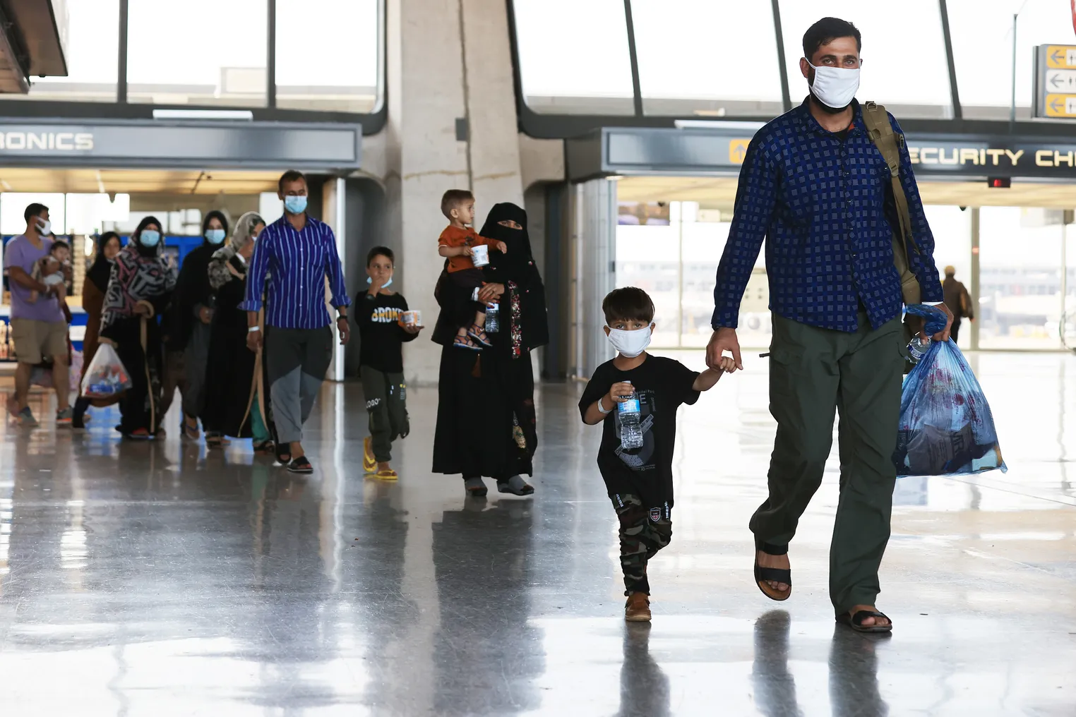 People of all ages arrive at an airport wearing masks and carrying bags and water bottles.