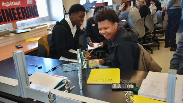 A student is sitting at a table in a classroom with other students. A metal gear device is sitting on the table
