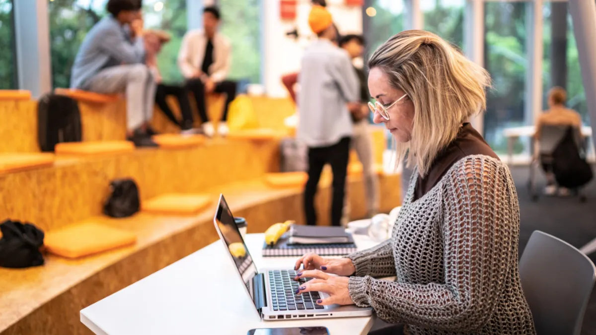 A person sitting at a table typing on a laptop with people blurred in the background