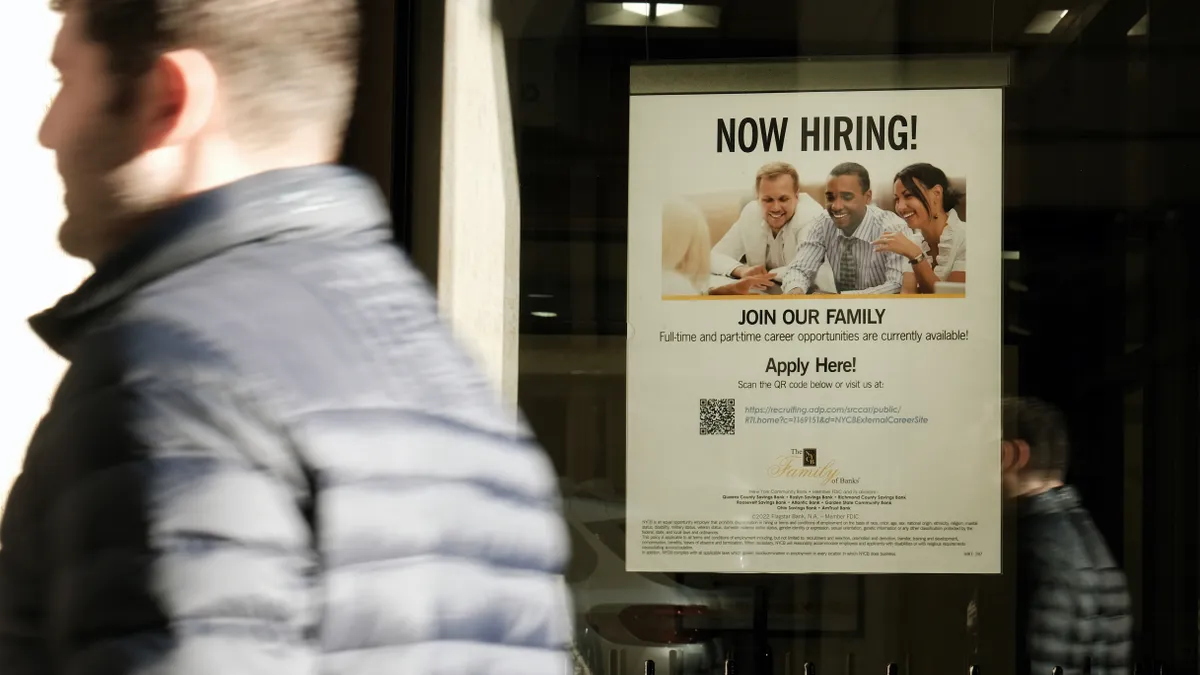 A person walk past a 'now hiring' sign is displayed in a store window.