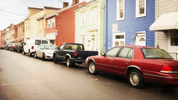 A line of cars parked in front of a sidewalk and houses.