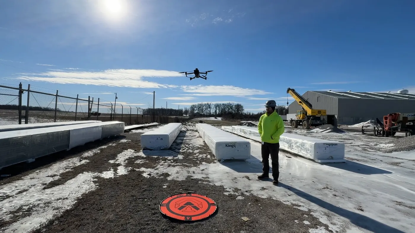 A person with a hard hat and a bright hoodie stands in a snow-covered landscape, while an aerial drone floats nearby.