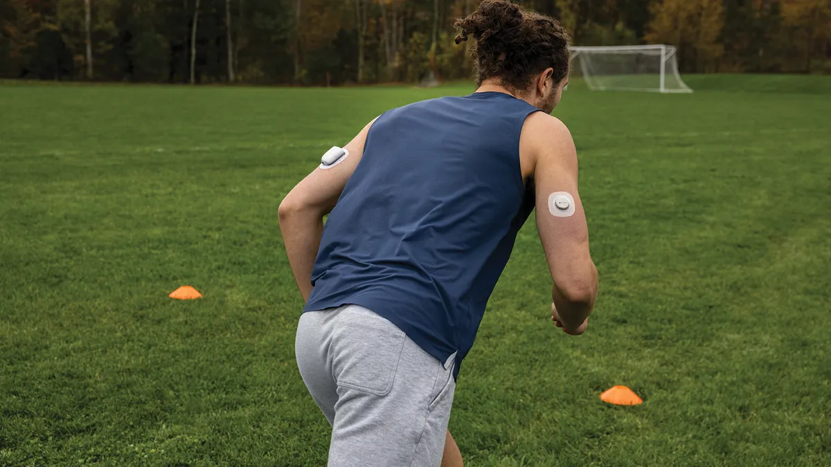 A person runs in a soccer field wearing two white devices taped to their upper arms.