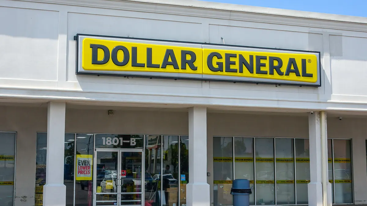 A plain white strip mall building with large glass windows. A bold yellow sign with black letters reads “DOLLAR GENERAL” above the entrance. The address “1801-B” is displayed on the door, and a trash bin is placed outside.
