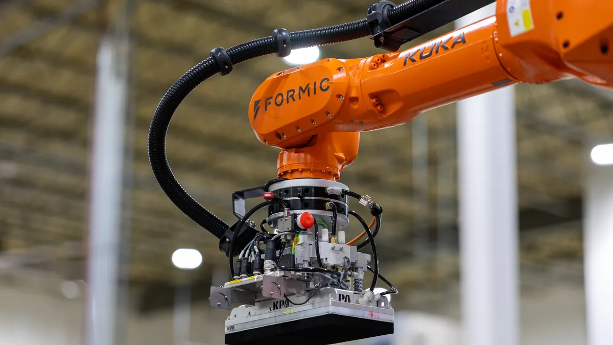 An industrial robotic arm labeled "FORMIC" and "KUKA" equipped with a complex gripper mechanism and wiring.