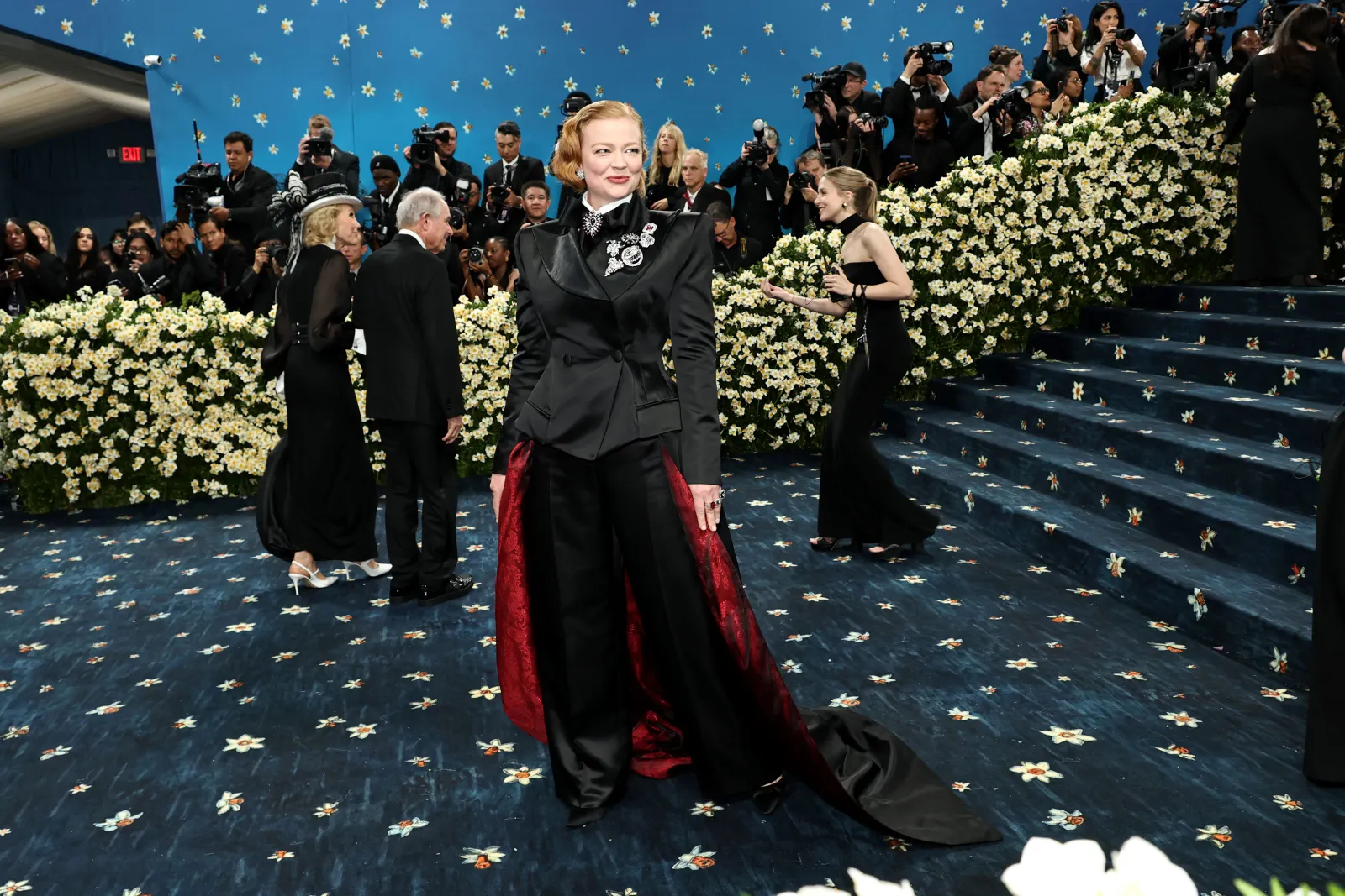 A woman in a black tuxedo and long topcoat with a train poses on a blue carpet.