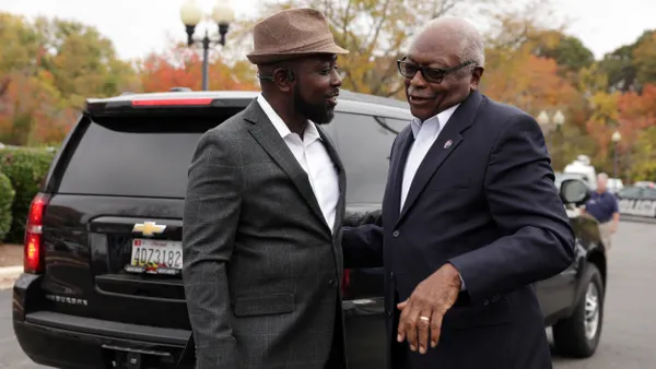 Two people wearing suits greet each other in front of a black Suburban.