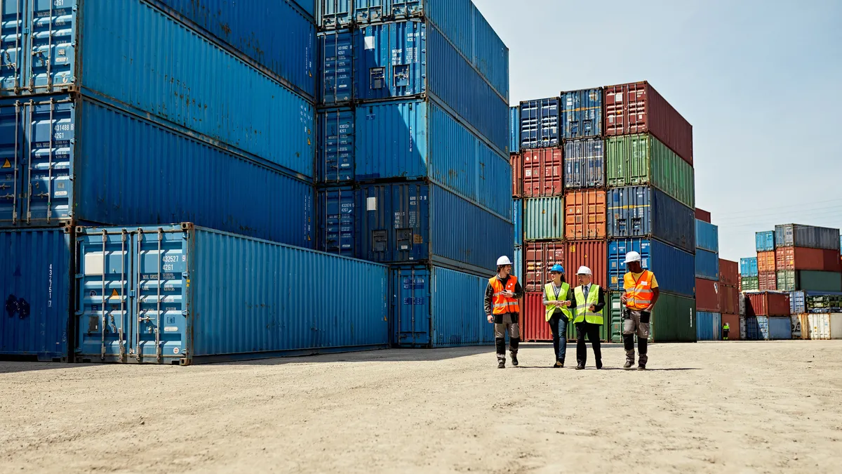 Mid distance view of diverse cargo handlers and supervisors approaching camera in reflective vests and hardhats amidst stacks of cargo containers.