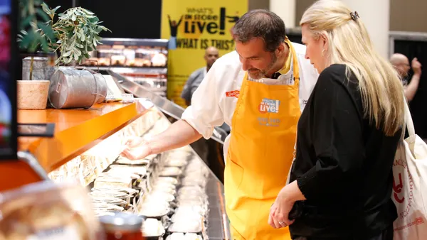 Person in an orange apron shows food products to someone at a grocery trade show.