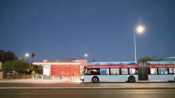A long blue bus on an empty road next to a bus shelter in late evening.
