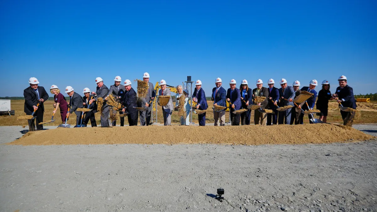 A line of 21 people across dirt, each wearing hard hats and carrying shovels, ceremonially break ground at the site of Novelis' aluminum rolling and recycling plant in Bay Minette, Alabama.