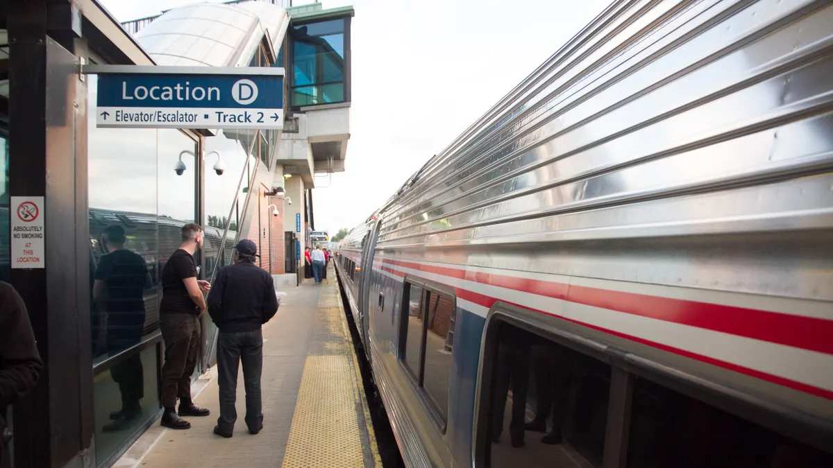 Two people in the foreground and more further back stand along a platform next to an Amtrak train.