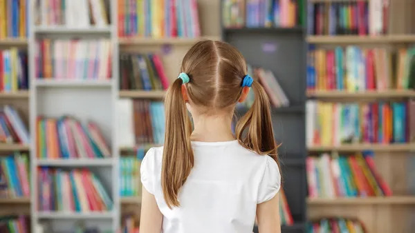 a young student with ponytails has their back to the camera and is facing a wall of shelves with multicolor books