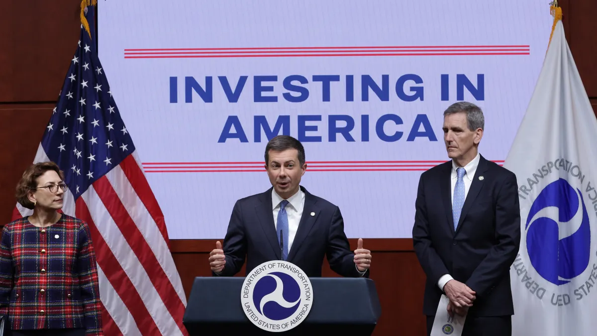 Man in suit at a podium in front of sign reading "Investing in America" with another man on the right and a woman on the left.