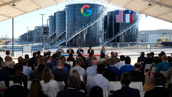 A panel of people sit at a table with a crowd sitting in chairs watching them in front of large round buildings with Google logo.