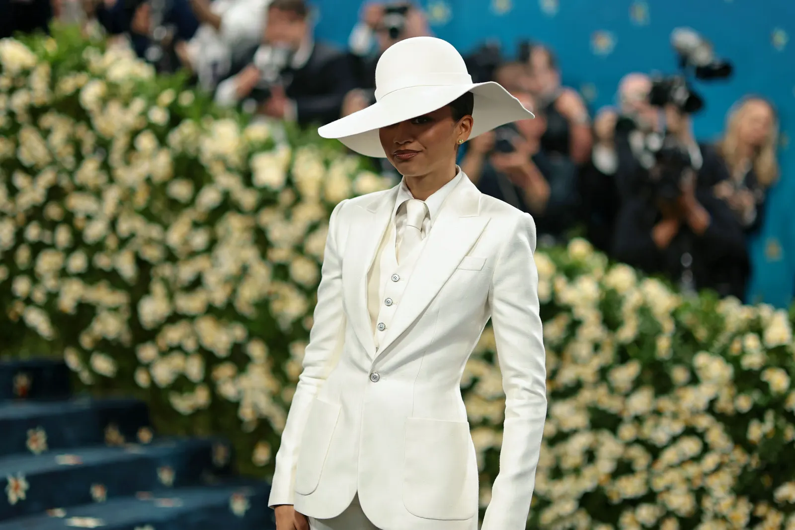 A woman stands wearing an all-white suit and white wide brimmed hat.