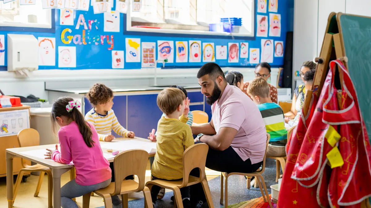 Adult sits a small table with younger students in a classroom and listens to one student talk.