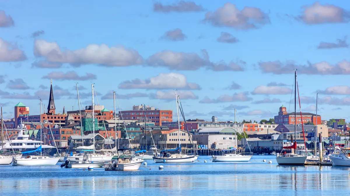 A shot of a bunch of sailboats in a bay on a sunny day. In the background, buildings rise above the horizon.