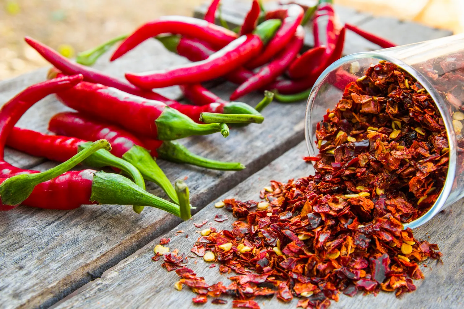 A photo of Red chili peppers and flakes on a wooden table outdoors. The flakes are spilling from a glass container.