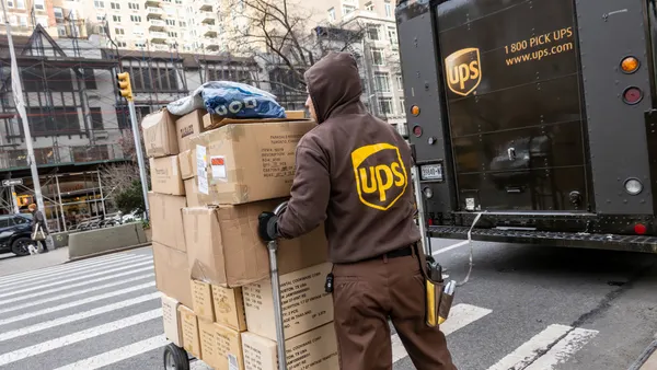 A person in a UPS uniform moves a cart of packages on the street in front of a UPS vehicle.