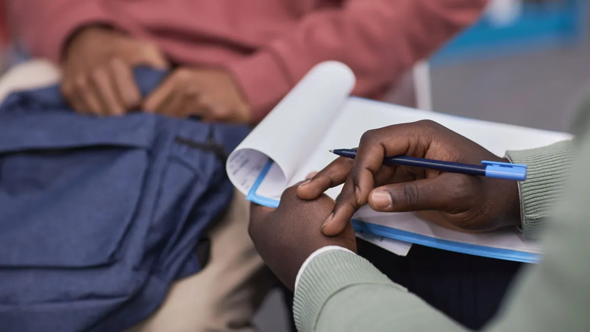 A closeup view of a counselor's hands holding a clipboard while listening to a student who is holding a backpack in their lap.