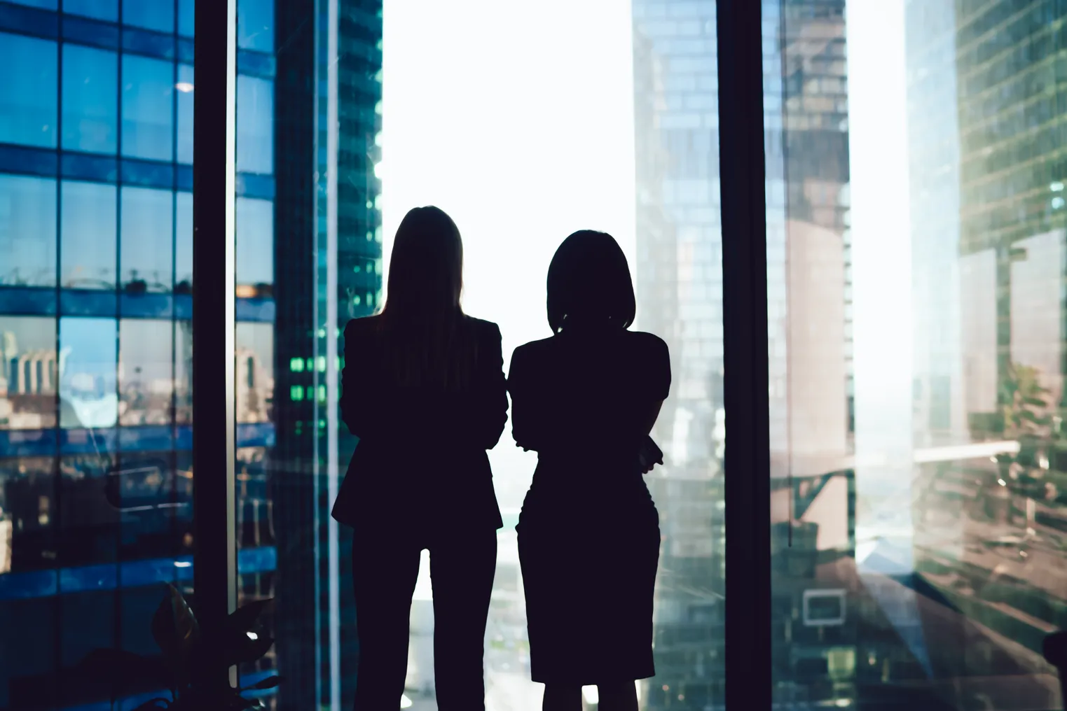 Back view of female colleagues in formal wear standing near window looking at modern exterior of skyscrapers in business center.