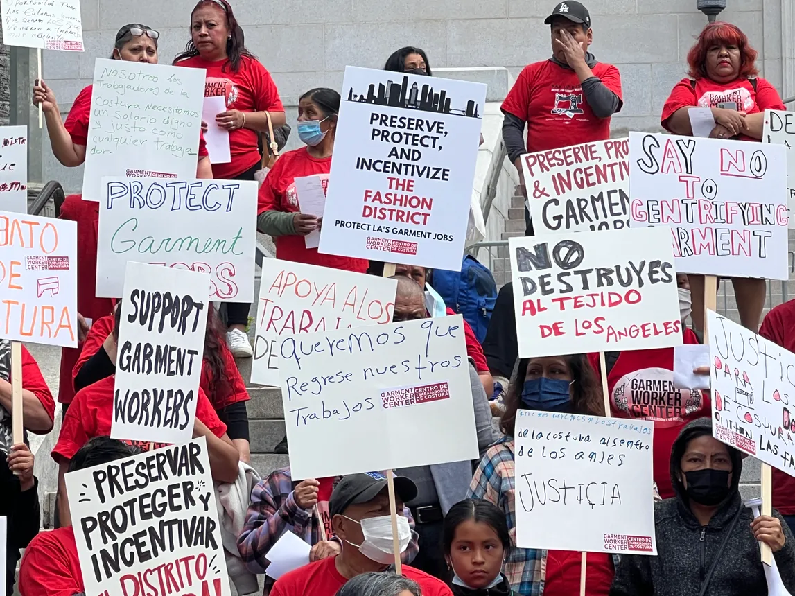 A crowd of people in matching red t-shirts stand in front of city hall carrying signs that read "support garment workers"