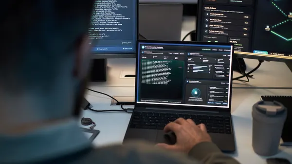 Caucasian person monitors cybersecurity dashboards across laptop and multiple screens, analyzing network threats and system data.