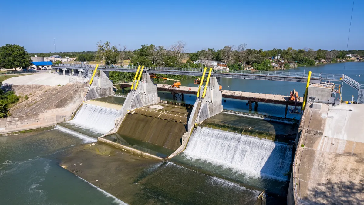 On a bright day, water flows over a dam and down into a larger body of water.