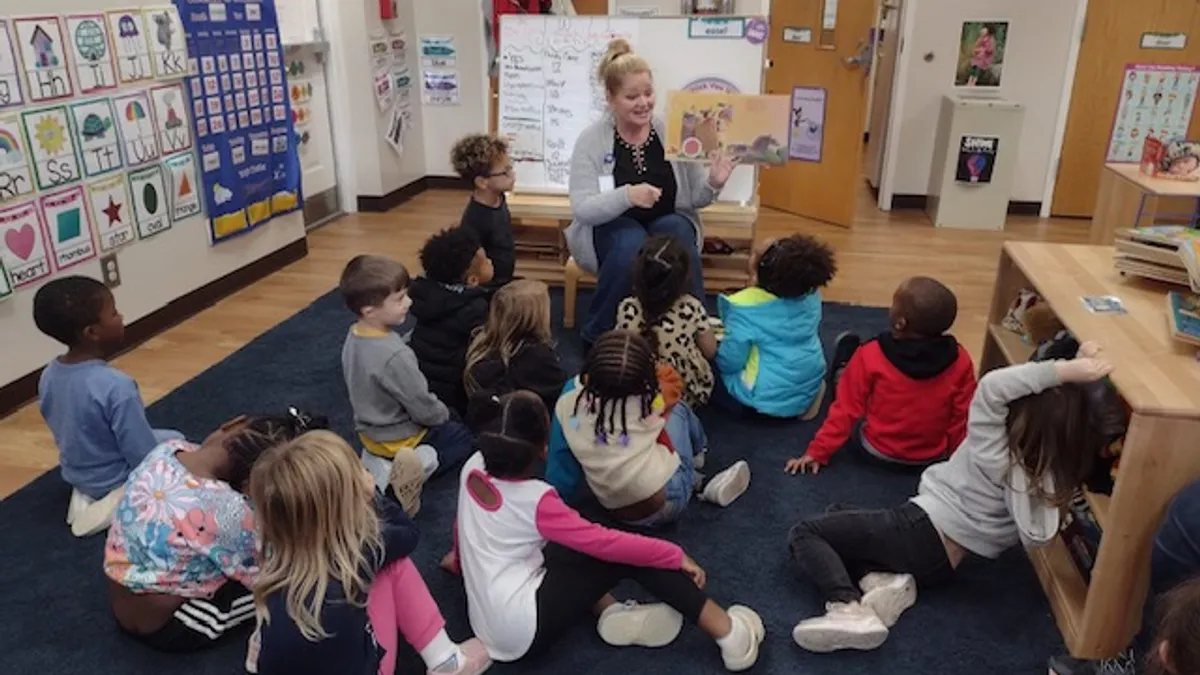 An adult teacher is in the front of a classroom sitting in a chair reading a book and showing the young students sitting in front of the teacher on the floor the pages in the book.