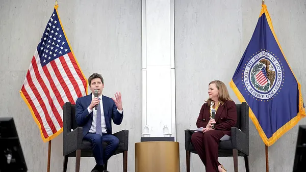 Sam Altman sits on the left side of a stage, in front of an American flag, while speaking into a microphone and gesturing, as Michelle Bowman sits on the right side of the stage in front of a Federal Reserve flag.