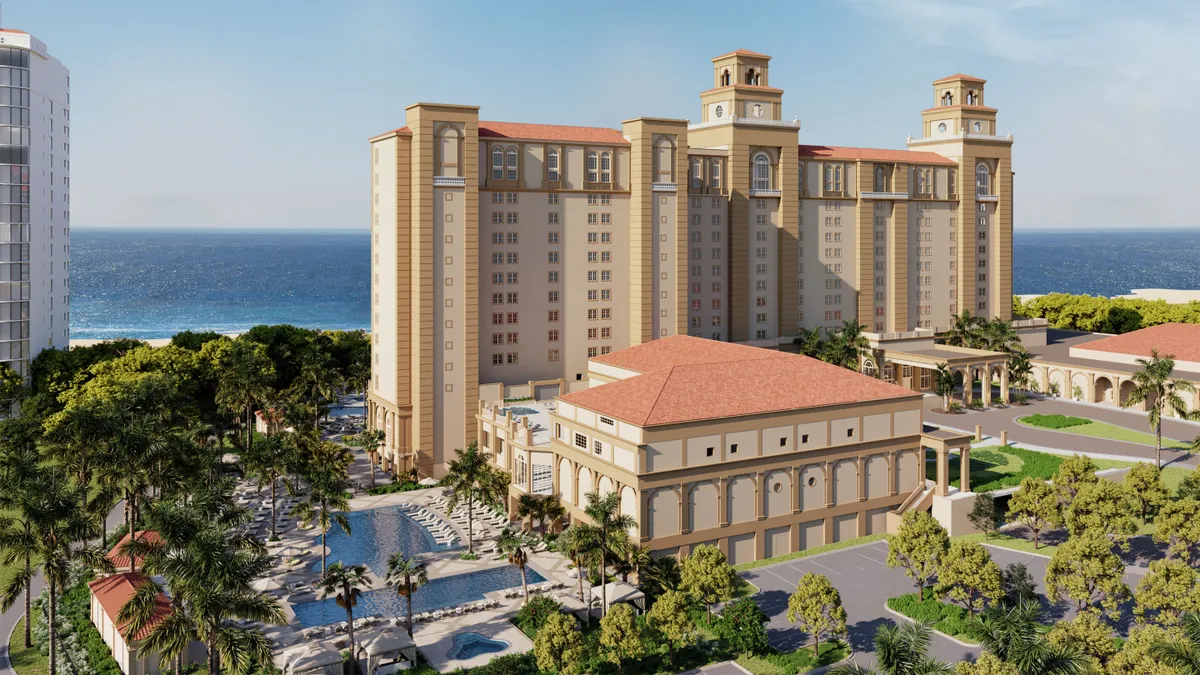 A hotel looms in the foreground on a bright, sunny day in Naples, Florida. The building is a beige stone with a reddish roofing, and the building is massive. The ocean is in the background.