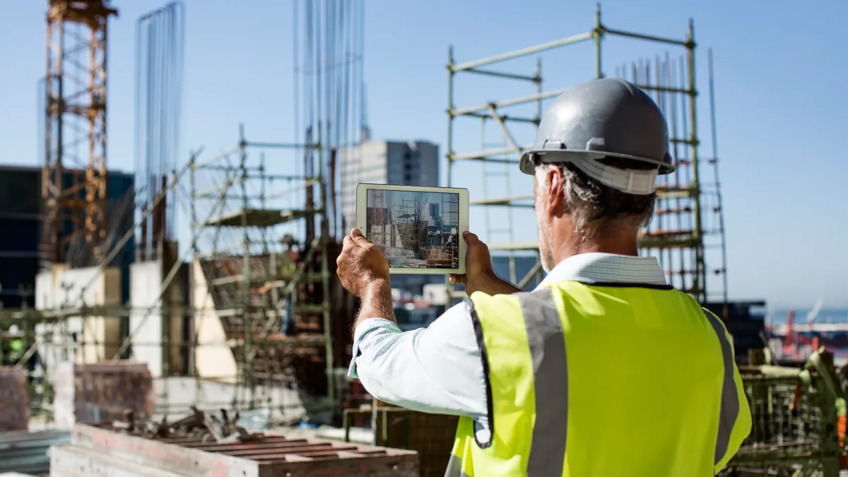 A person in safety gear, which includes a reflective vest, hardhat and safety glasses, holds a tablet up to take a photo of a construction site.