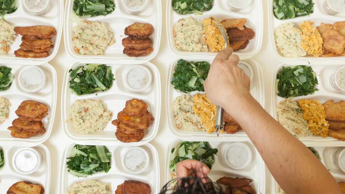 A view of a person's hand assembling multiple rows portioning meals to be served out on individual serving trays.