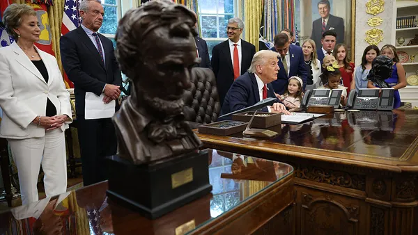 One person is seated at a desk while several adults and children are gathered around. A bust of Abraham Lincoln is in the foreground