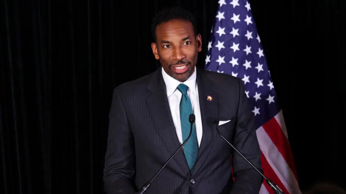 A person in a suit and ties stands behind a podium. Behind him is a United States flag.