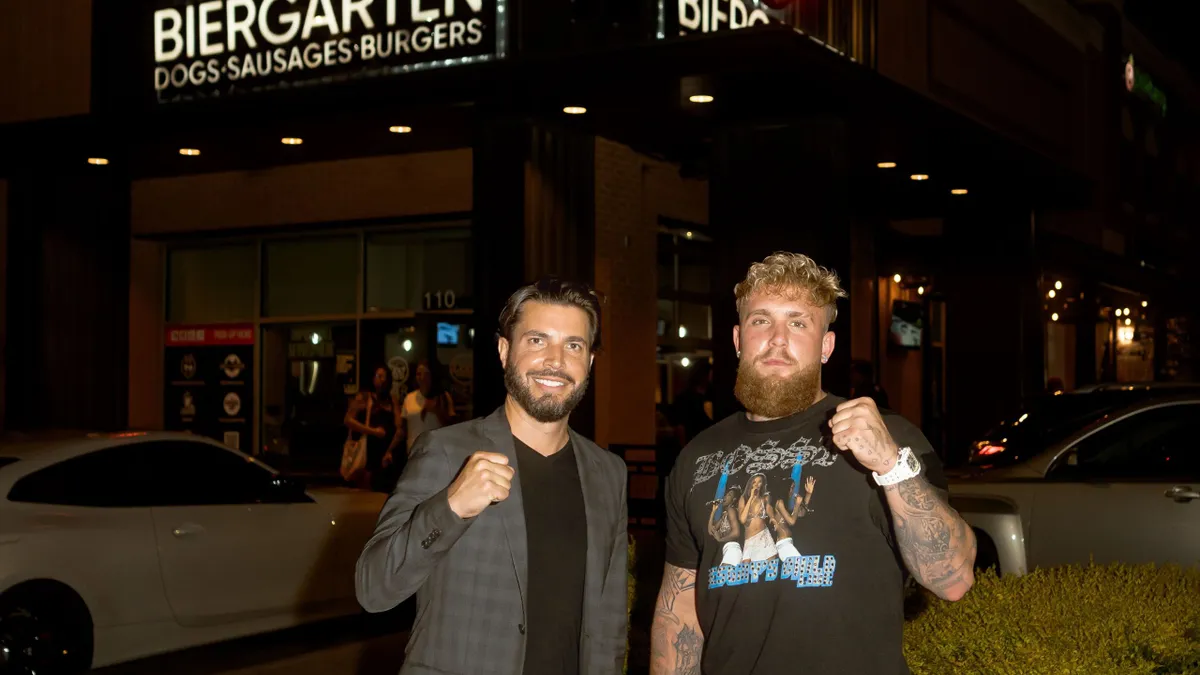 Michael Montagano, Dog Haus' CEO, and Jake Paul in front of a Dog Haus restaurant at night. Montagano wears slacks and a sport coat, Paul wears cargo shorts and a t-shirt.