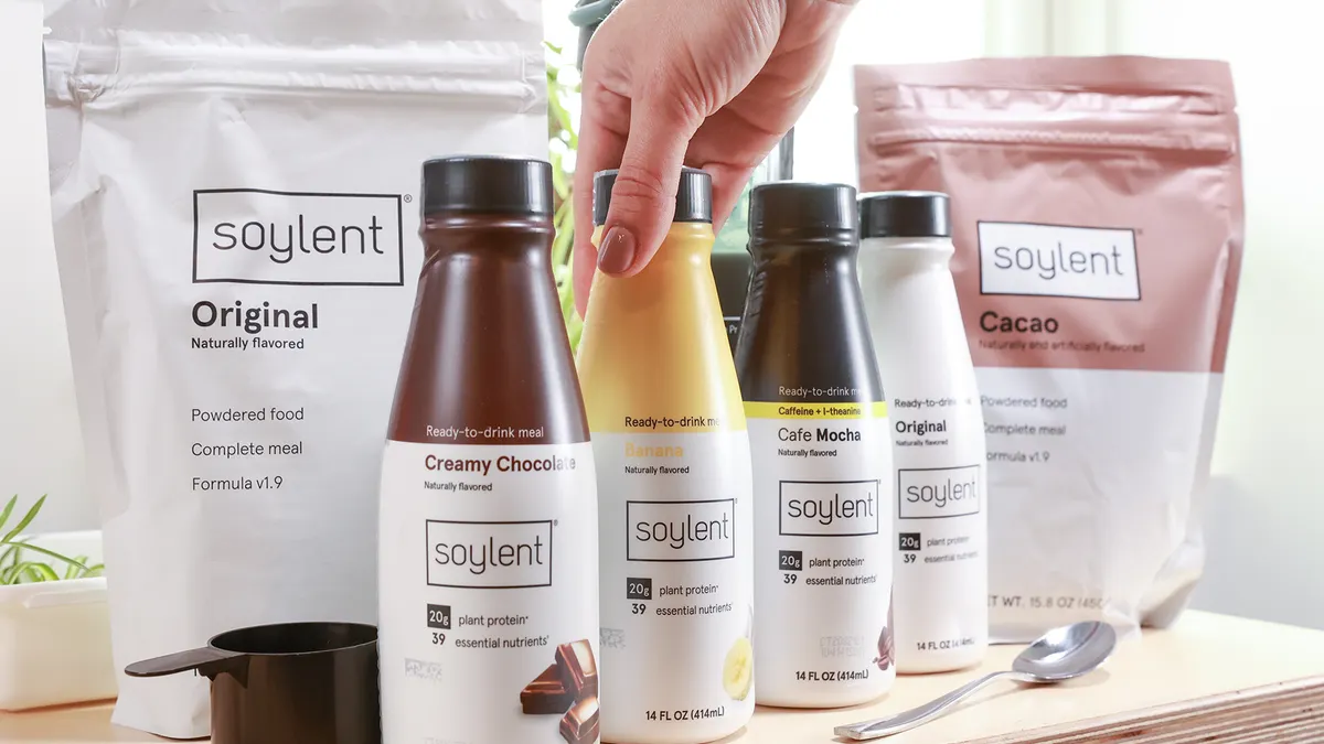 Two pouches of Soylent protein powder and three four different bottles of Soylent beverages sit on a table with a measuring cup and a spoon. A hand touches one of the bottles.