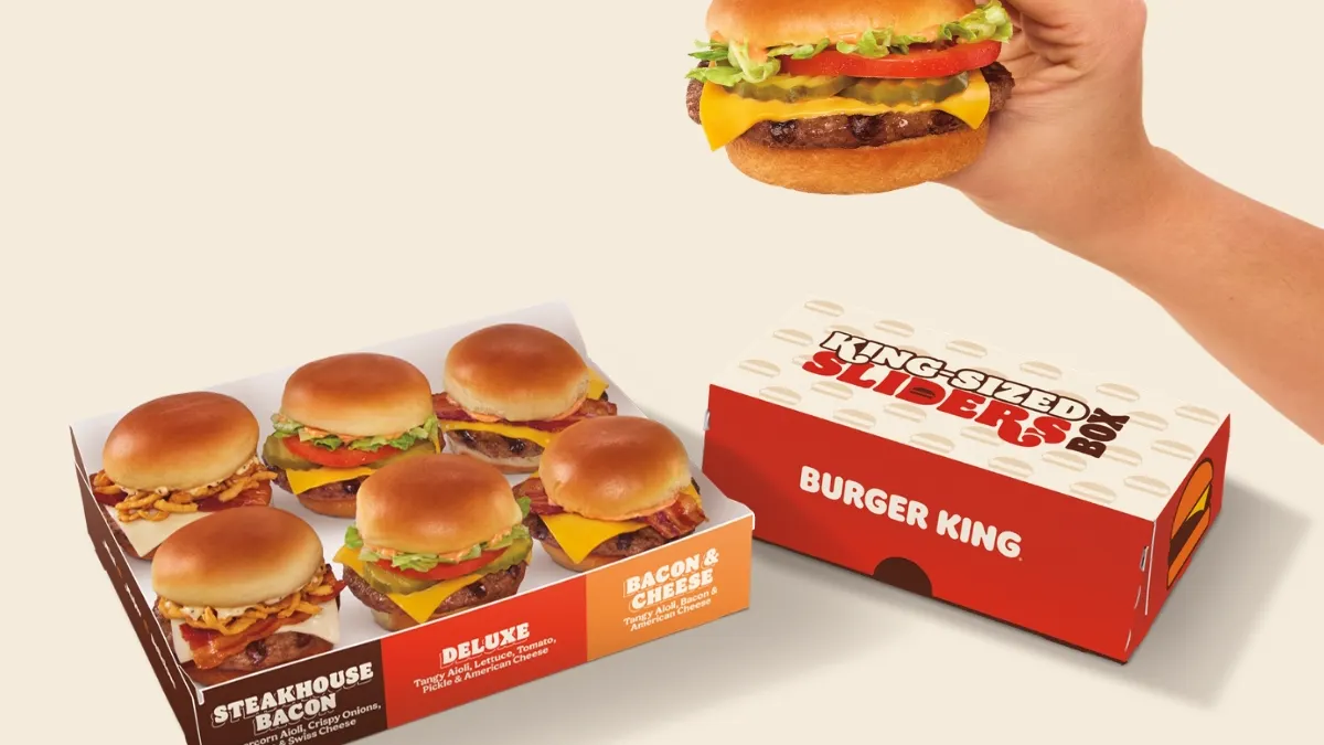 A hand holds a Burger King slider in front of boxes of similar miniature burgers.