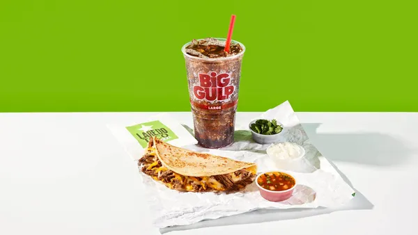 A photo of a tsco on a napkin surrounded by a cup of soda and several cuts of garnishes and dips. the napkin says Laredo Taco and the soda cup says Big Gulp.