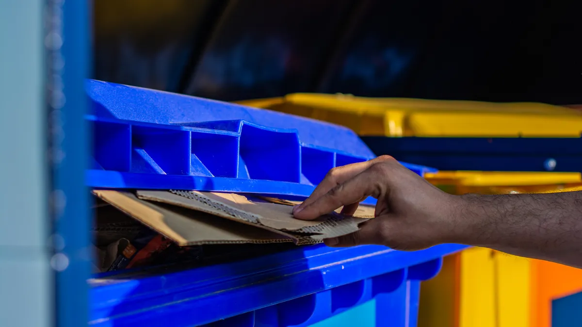 A person's hand inserts cardboard into a blue recycling cart