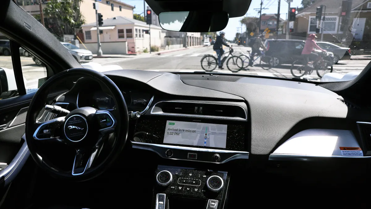 View from inside a vehicle  without a driver approaching an intersection with cars and bicycles.