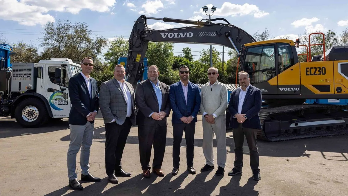 Six people in suits stand in front of an electric collection truck and excavator in Florida