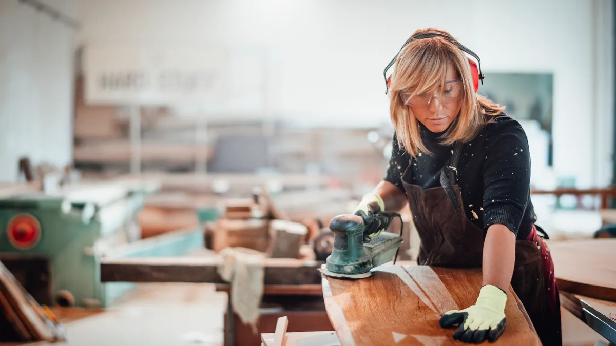 A feminine person uses a handsander to smooth out some medium-toned wood