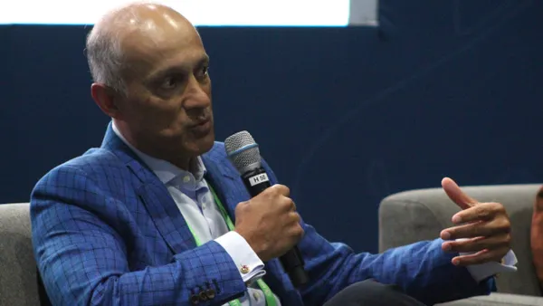 A person in a blue jacket holds a microphone and gestures as they speak.