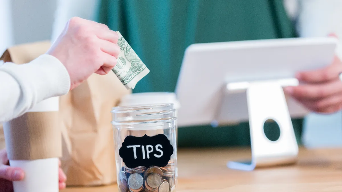 An image of a hand putting a dollar in a tip jar.