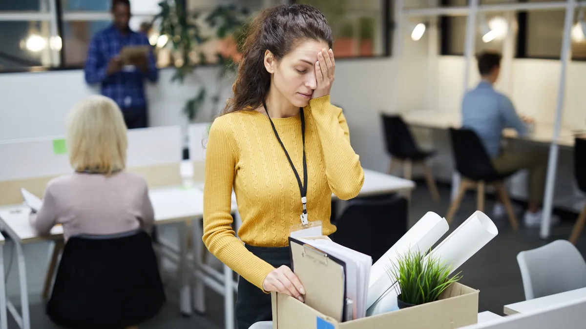 A frustrated young person in yellow sweater stands at table and touches their face with their hand while packing stuff up in office after dismissal