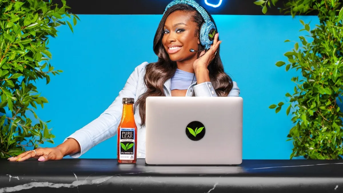 Actress-singer Coco Jones wears a headset behind a laptop in front of a neon sign that reads "Subtly Sweet Hotline"