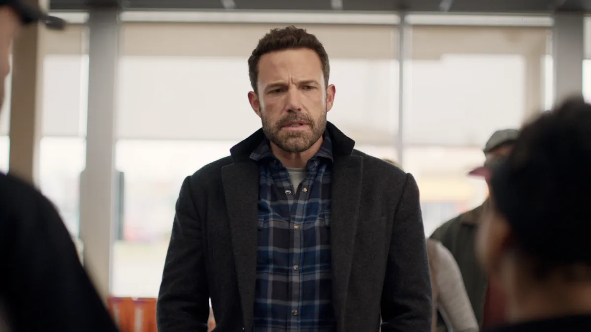 Actor Ben Affleck, wearing a blue plaid shirt and dark coat, stands center frame in front of a Dunkin' cash register.