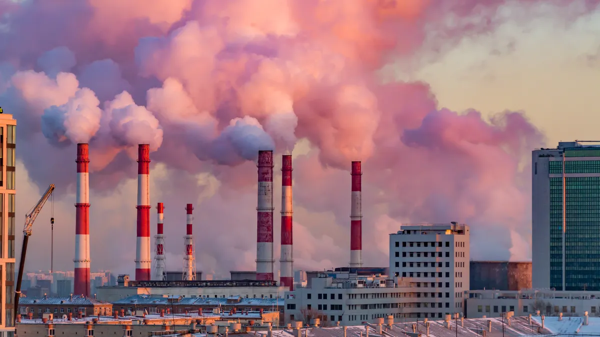 Steam rises from factory pipes in a city and the sky behind the pipes is bright pink and orange.